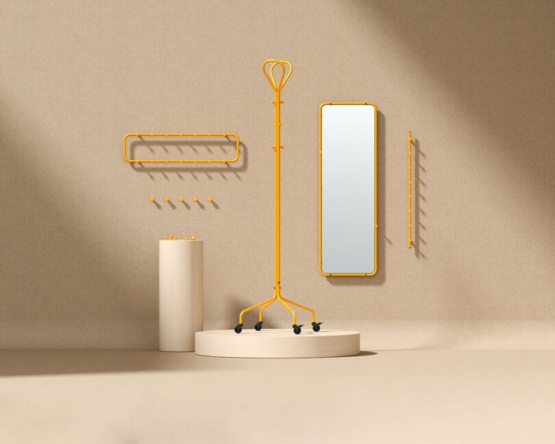 A minimalist display features yellow metal coat racks, hooks, and a tall mirror on a beige background with a round pedestal and a cylindrical stand.