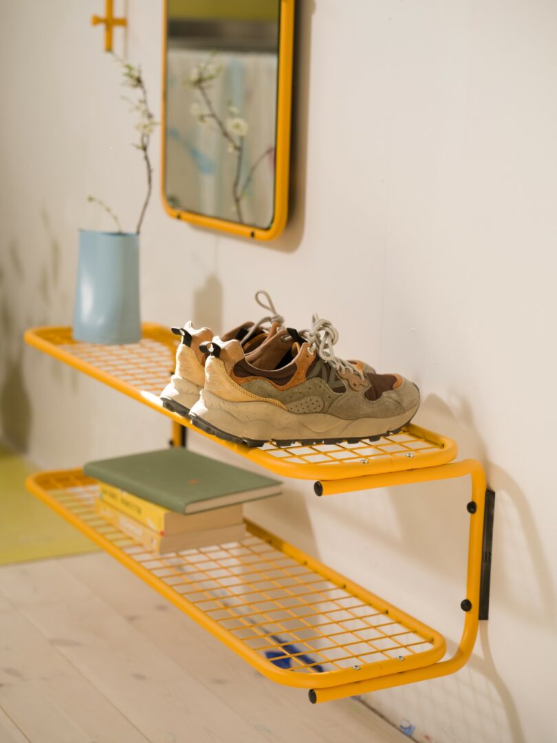A pair of sneakers sits on the top yellow wire shelf, with books and a blue vase on the lower shelf, against a light-colored wall with a mirror