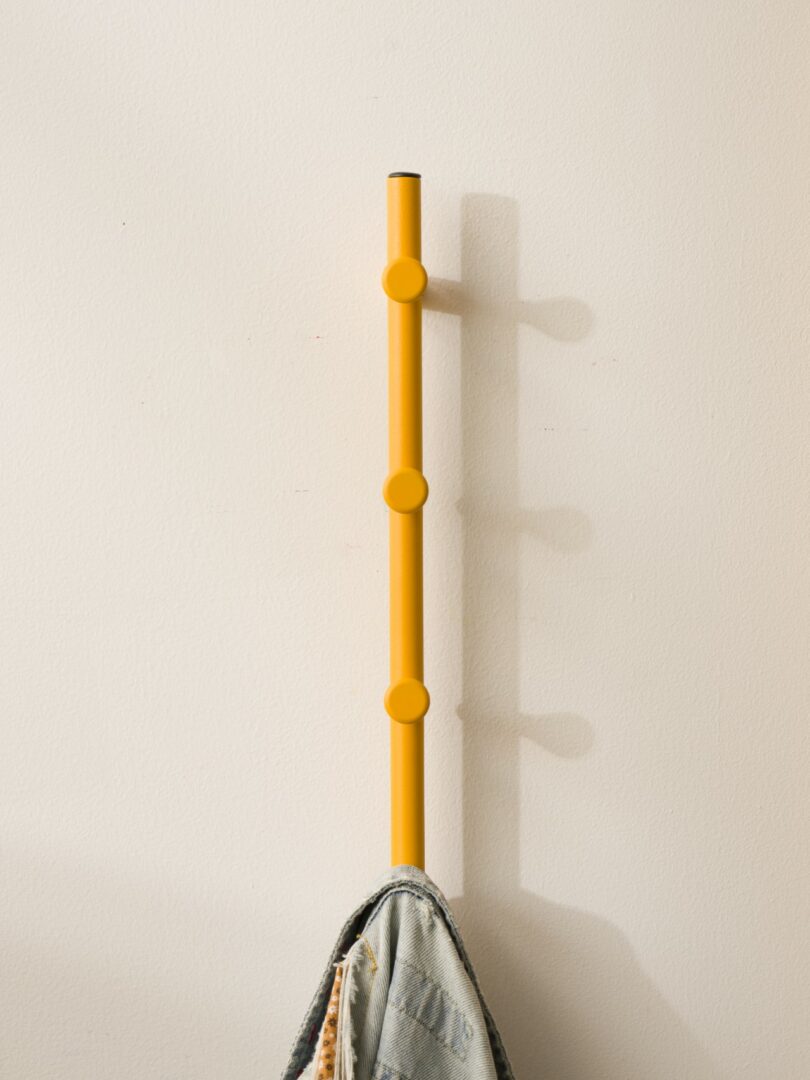 A yellow wall hook rack with three round pegs is mounted vertically on a light-colored wall, holding a piece of clothing at the bottom peg