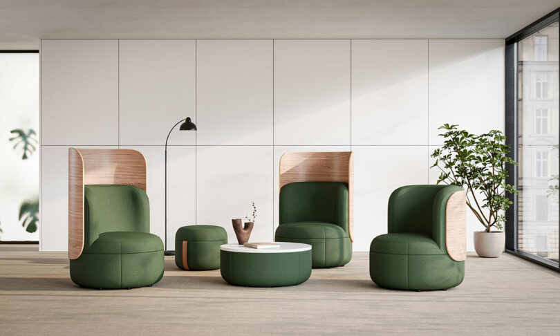 Modern office lounge with green upholstered chairs, a round white coffee table, a floor lamp, and a potted plant, set against a white paneled wall and large windows.