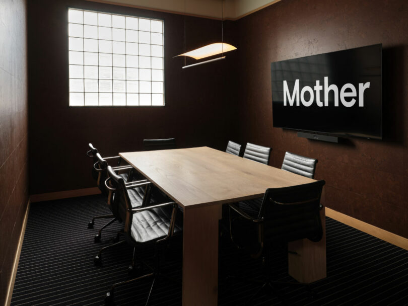 A conference room with a rectangular table, seven black chairs, a large window, and a wall-mounted screen displaying the word 