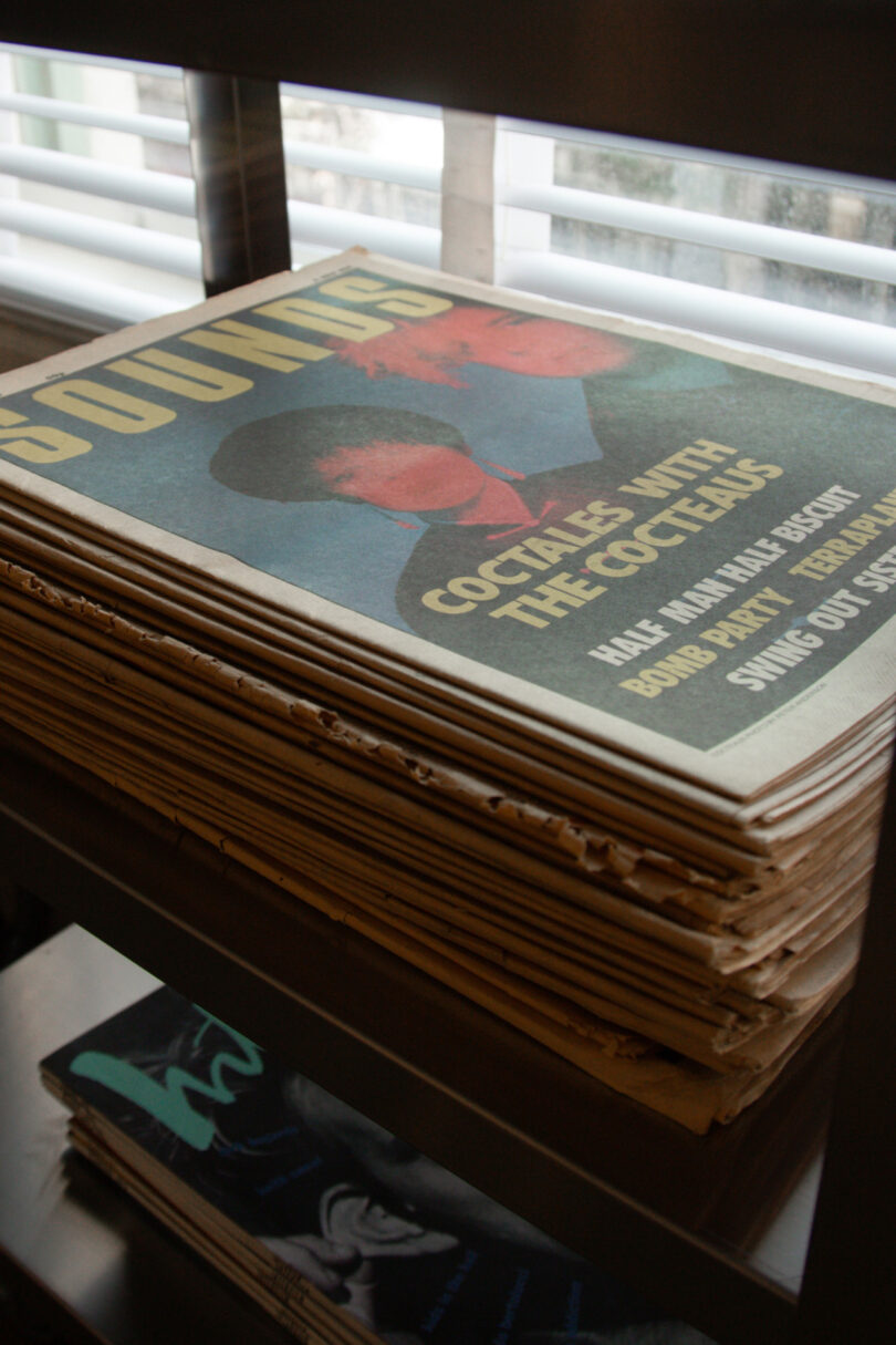 A stack of vintage magazines sits on a metal shelf near a window, with the top issue featuring a cover story titled