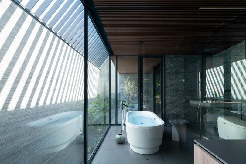 Modern bathroom with a freestanding bathtub next to large floor-to-ceiling windows, featuring natural light and diagonal shadow patterns on the floor and walls.