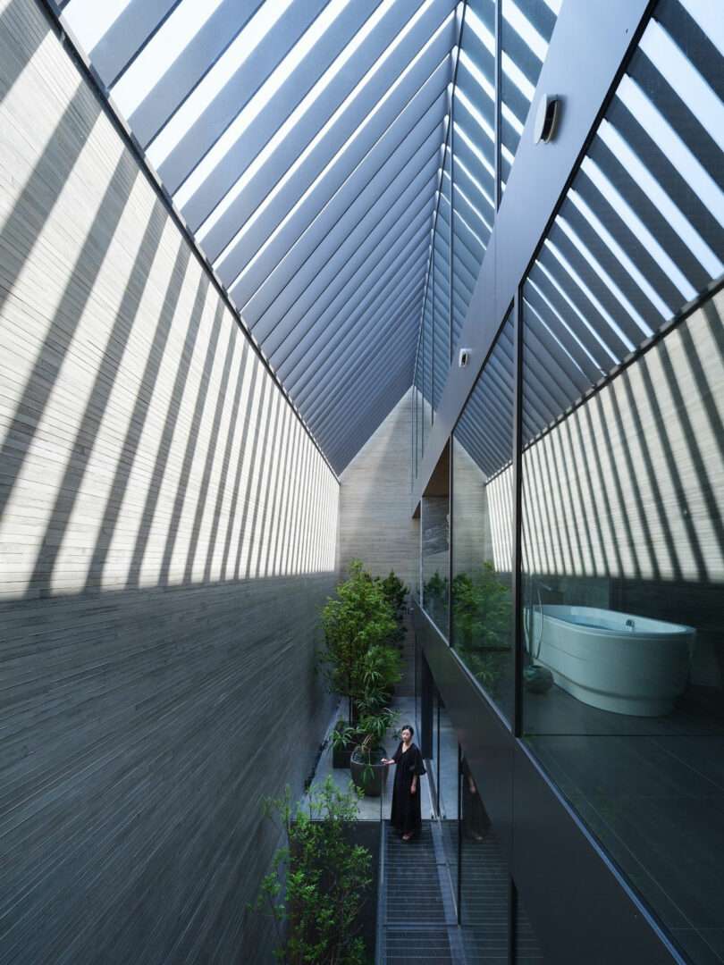 A person stands in a narrow, modern corridor with a slanted glass roof casting shadows, next to a glass wall revealing a bathtub and indoor plants.