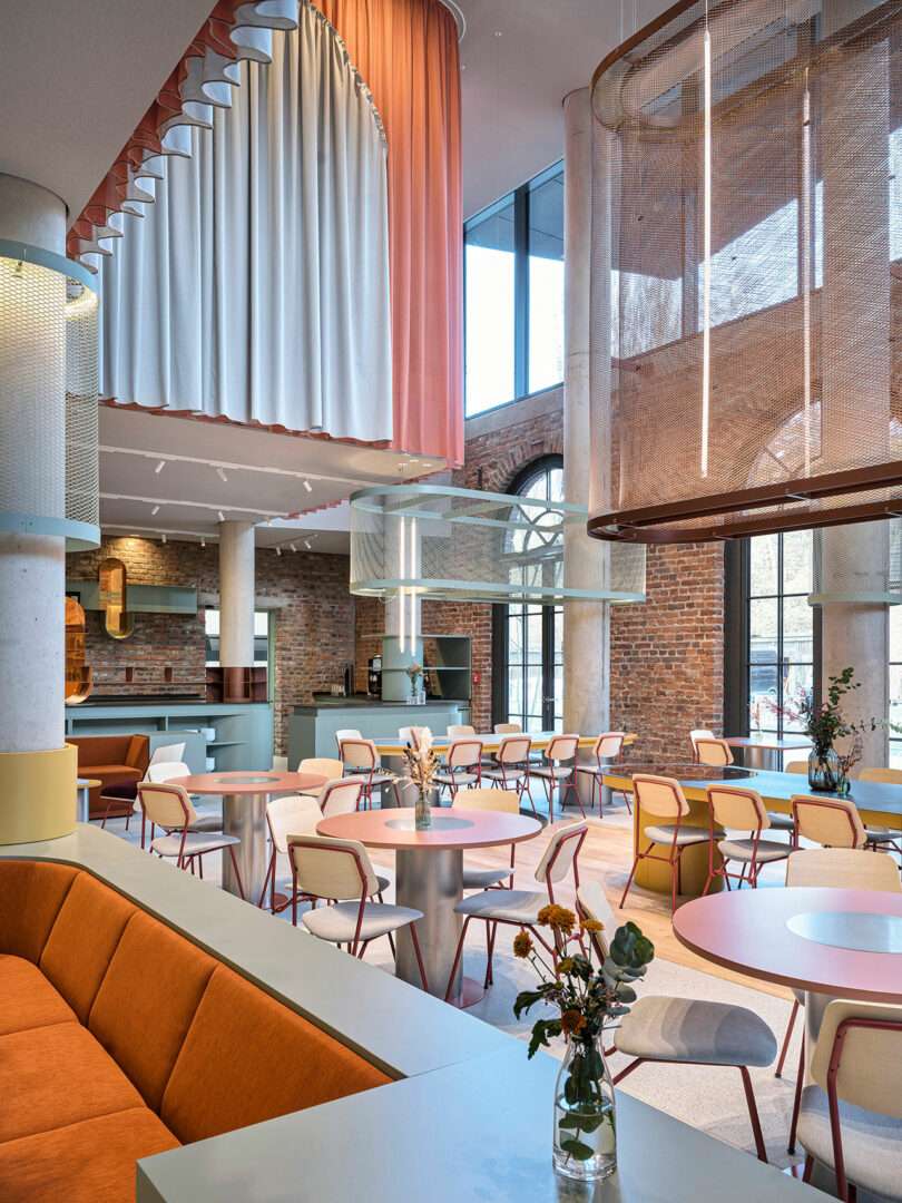 Modern café interior at NOXX Hotel features brick walls, large windows, pastel furniture, round tables, orange sofa seating, and decorative hanging panels. A vase with flowers sits on a table in the foreground.
