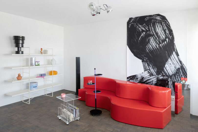 Modern living room with a red modular sofa, abstract black-and-white wall art, minimalist shelving with decor items, and geometric accessories on a wooden floor.