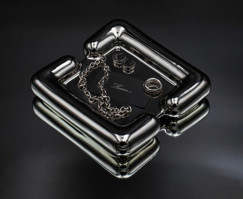 A metallic tray holds a chain necklace, two rings, two cylindrical objects, and a card labeled
