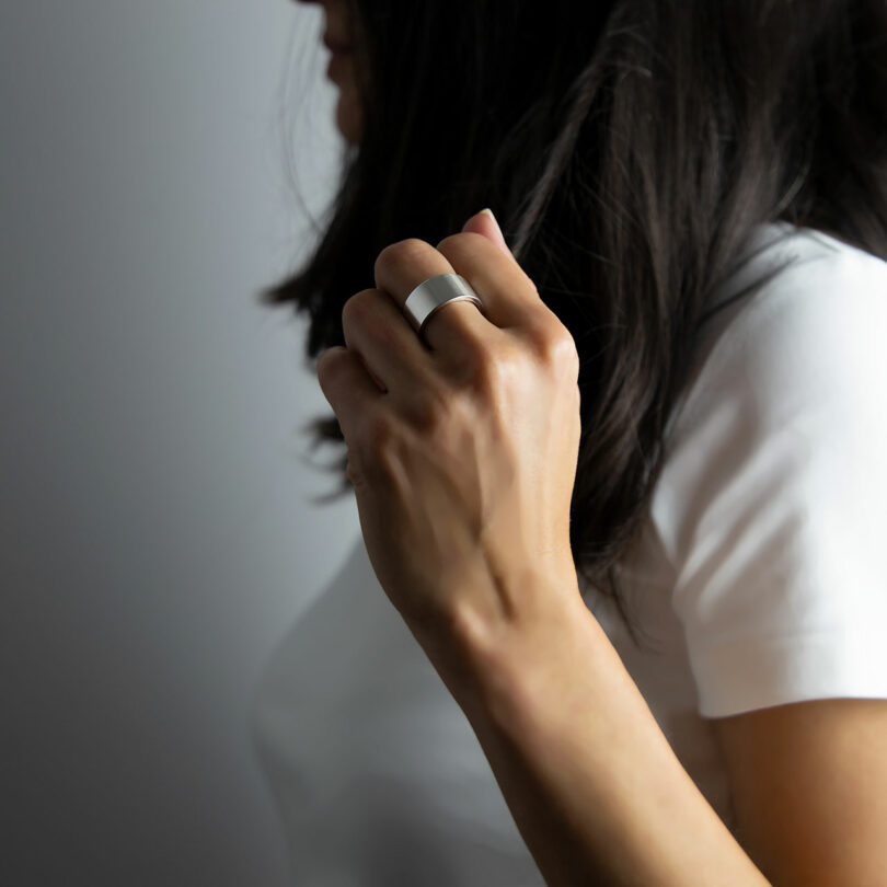 A person with long dark hair wearing a white shirt holds up their hand, displaying a wide silver ring on their finger against a plain background.