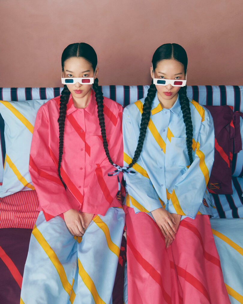 Two women with long braids sit side by side on a striped bed, wearing colorful, matching patterned outfits and retro 3D glasses.