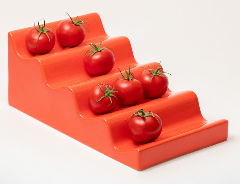 Six tomatoes arranged on a bright orange, stepped display stand, against a plain light background.