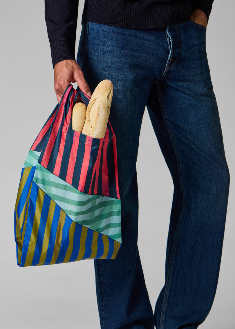 A person in blue jeans holds a multicolored striped reusable bag with two baguettes partially visible inside.