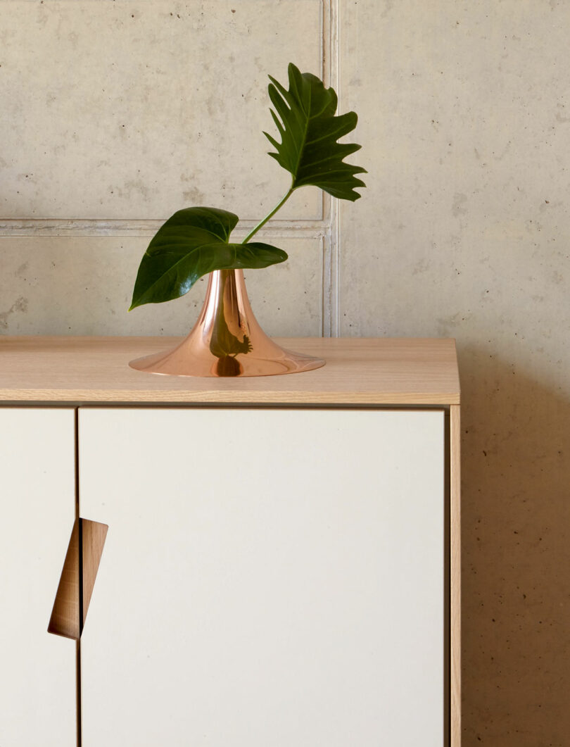 A single green leaf in a modern copper vase sits atop a light wood cabinet against a neutral textured wall—an elegant touch perfect for any Gift Guide.
