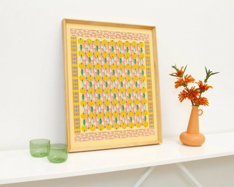 A framed geometric art print, two green glass cups, and an orange vase with orange flowers are arranged on a white shelf against a plain white wall.