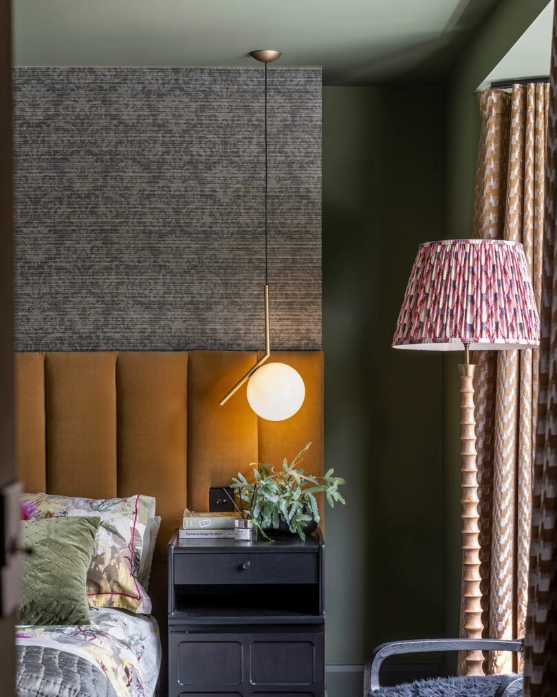 A modern bedroom with a tan upholstered headboard, a round pendant light, a black nightstand with books and a plant, and a pleated lampshade floor lamp.