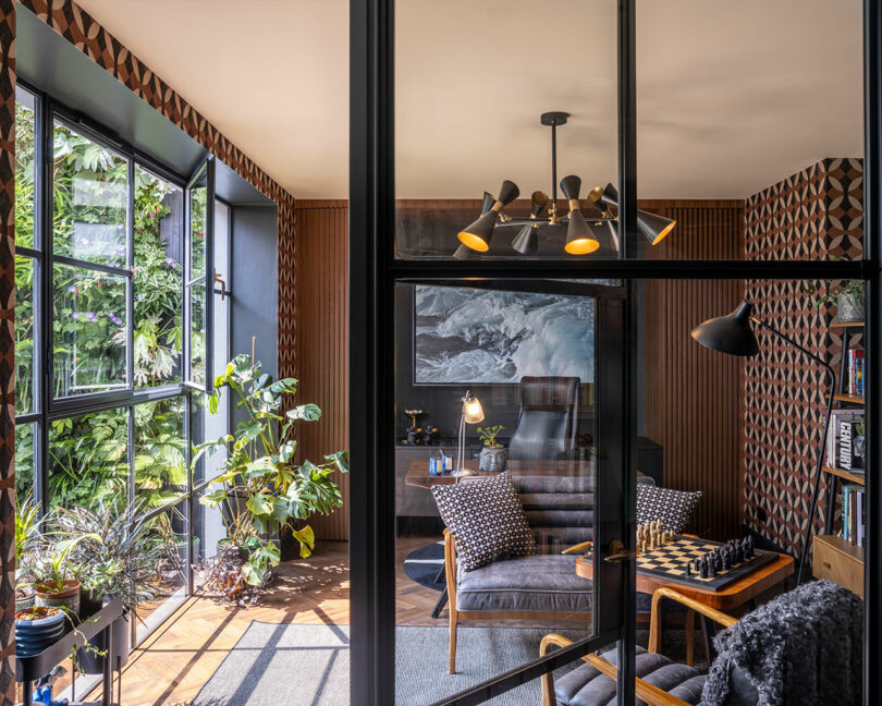 Modern home office with glass doors, patterned walls, indoor plants, a desk, bookshelves, a chessboard on a small table, and large windows letting in natural light.