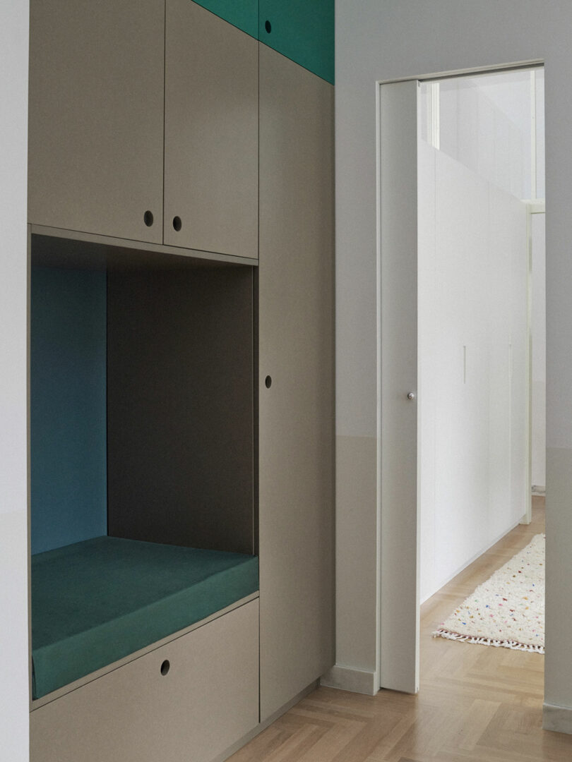 Built-in storage cabinets with a recessed bench and teal accents beside an open doorway leading to a hallway with white walls and a patterned rug.