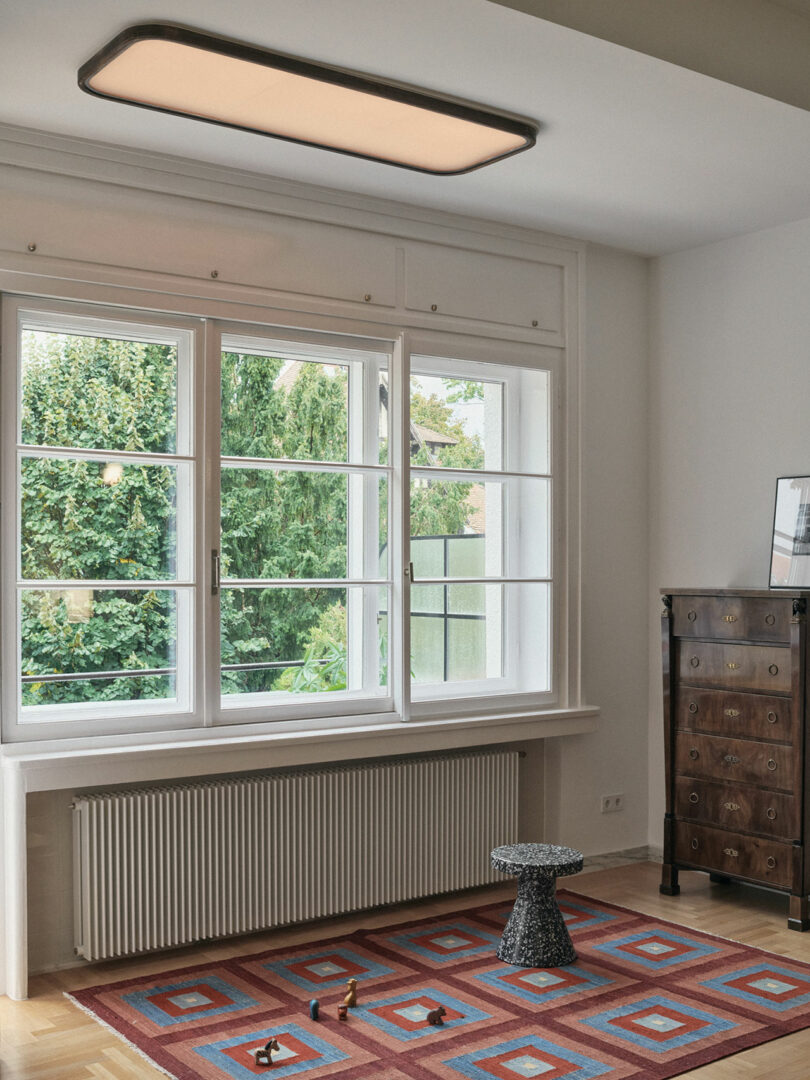 A bright room with a large window, patterned rug, small textured table, wooden dresser, and a few small toy animals on the floor.