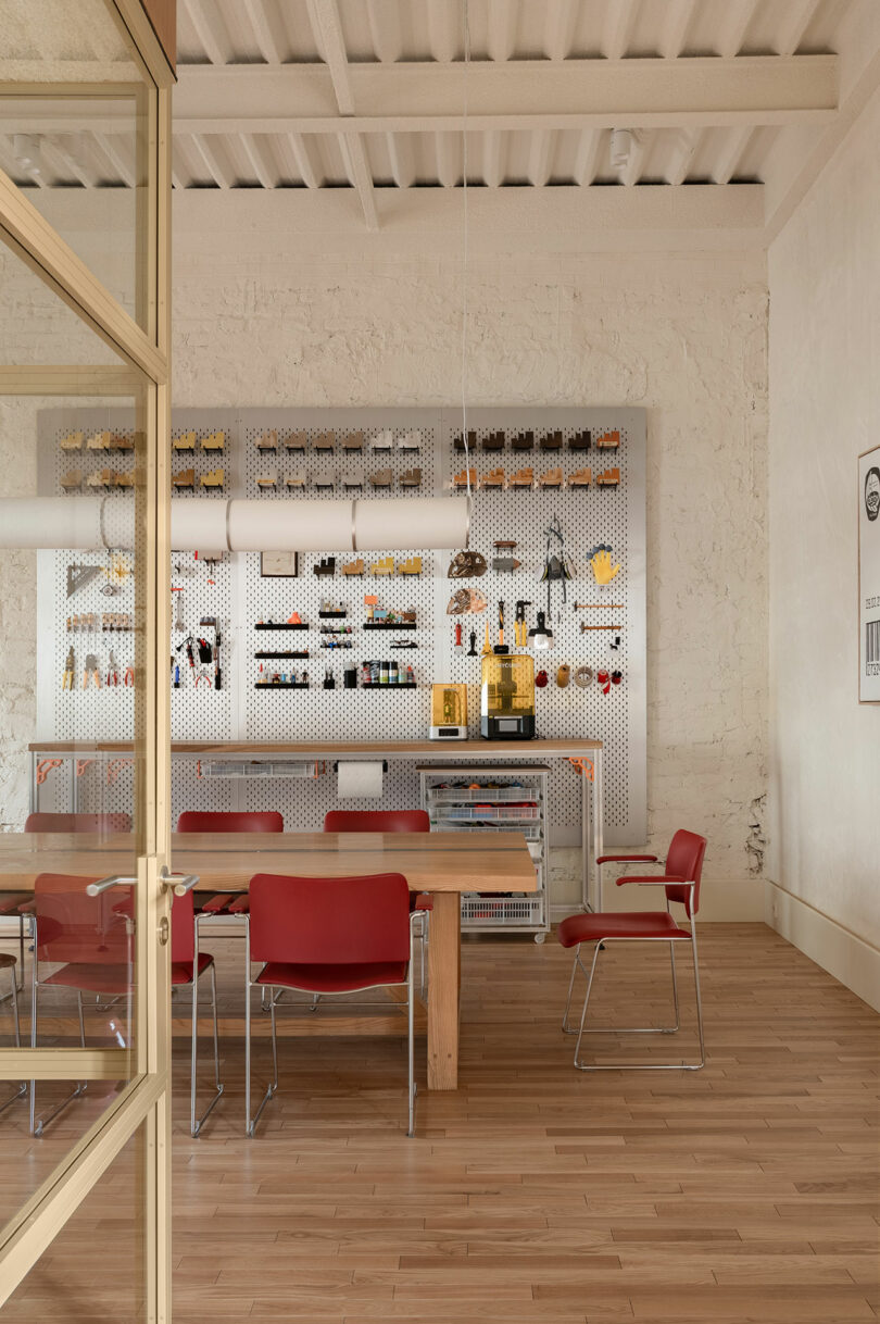 A modern meeting room with a wooden table, red chairs, and a pegboard shelf holding various office supplies and decorative items against a white brick wall.