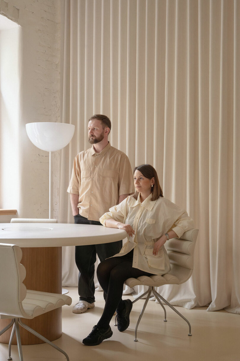 A man stands next to a woman seated at a round white table in a minimalist room with light-colored curtains and modern chairs.