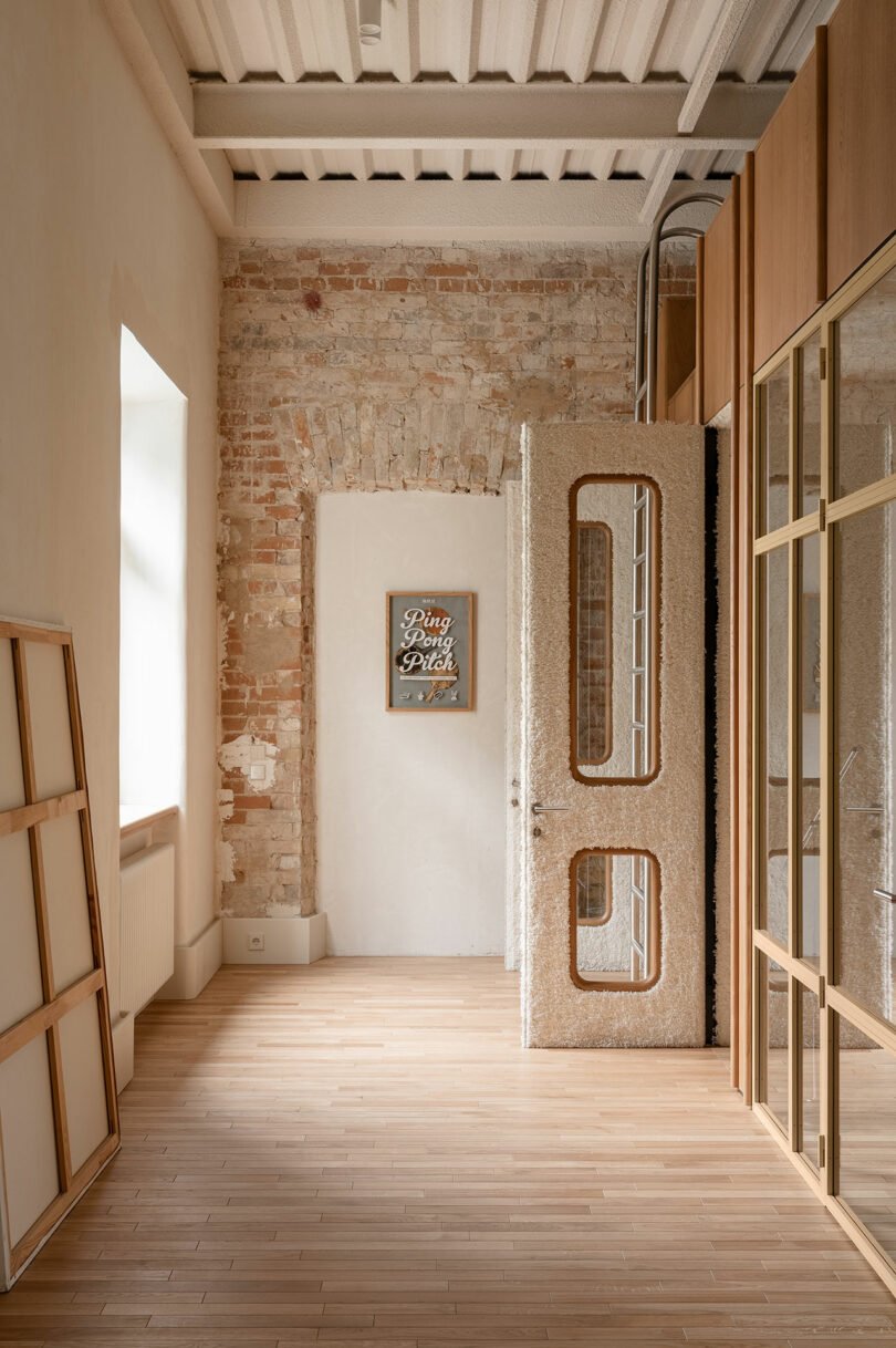 A bright hallway with exposed brick and plaster walls, a textured glass door, wooden floor, and a framed artwork that reads 