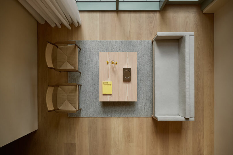 A minimalist living room viewed from above, featuring two chairs, a sofa, a wooden coffee table with a book and flowers, all set on a light gray rug over wooden flooring.