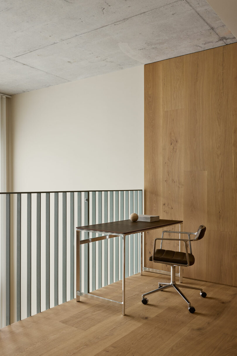 A minimalist workspace with a metal-framed desk, brown chair, and a ball and book on the desk, set against wooden and concrete walls beside a metal railing.