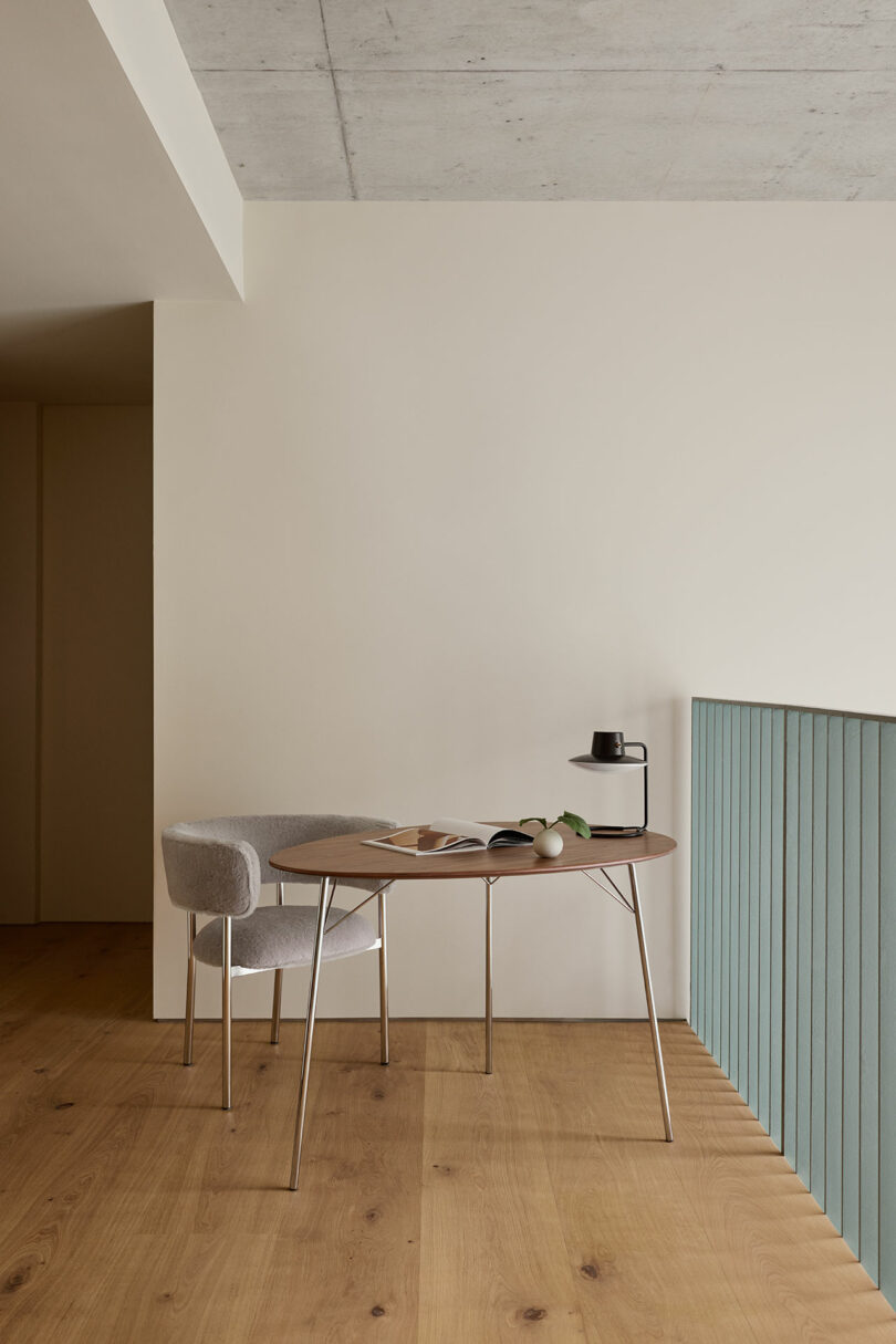 A minimalist workspace with a round table, gray chair, open book, potted plant, and black carafe on light wood flooring next to a pale green railing.