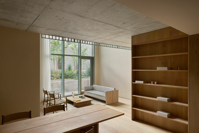 Minimalist living space with large window, light wood furniture, built-in shelves, and a view of a small terrace garden outside.
