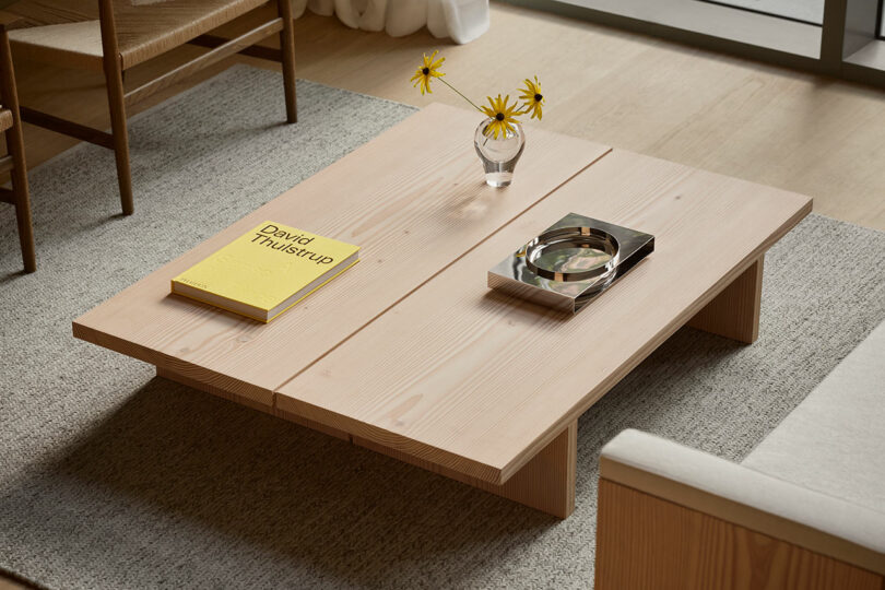 A light wood coffee table with a yellow book, a glass vase with yellow flowers, and a metal tray, set on a gray rug in a modern living room.