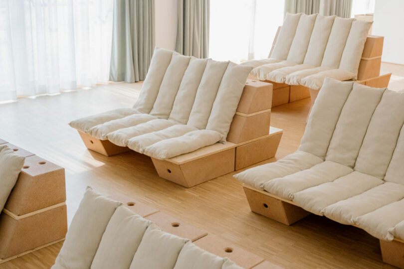 Several cushioned lounge chairs with wooden bases are arranged in a sunlit room with light wood flooring and large windows covered by sheer curtains