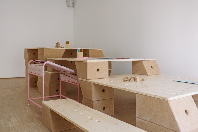 A minimalistic room with modular wooden tables and benches, cork block supports, a pink bench, and small scattered objects on the surfaces