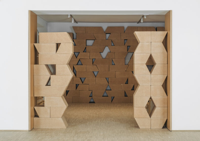 A room features a sculptural partition wall and doorway made of interlocking geometric cork blocks with hexagonal cutouts, set against a plain interior backdrop