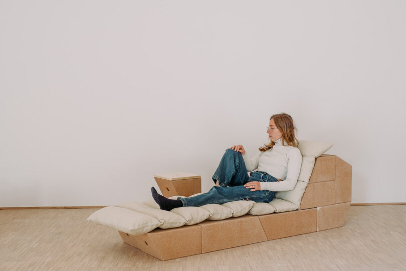 A person sits on a modular, cushioned lounge chair made of wooden panels with a pillow, next to a small box-shaped side table in a minimalist room