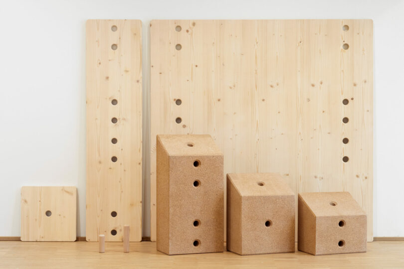 Five rectangular wooden boards with circular holes are arranged vertically behind four cork blocks, also with holes, on a light wood floor against a white wall