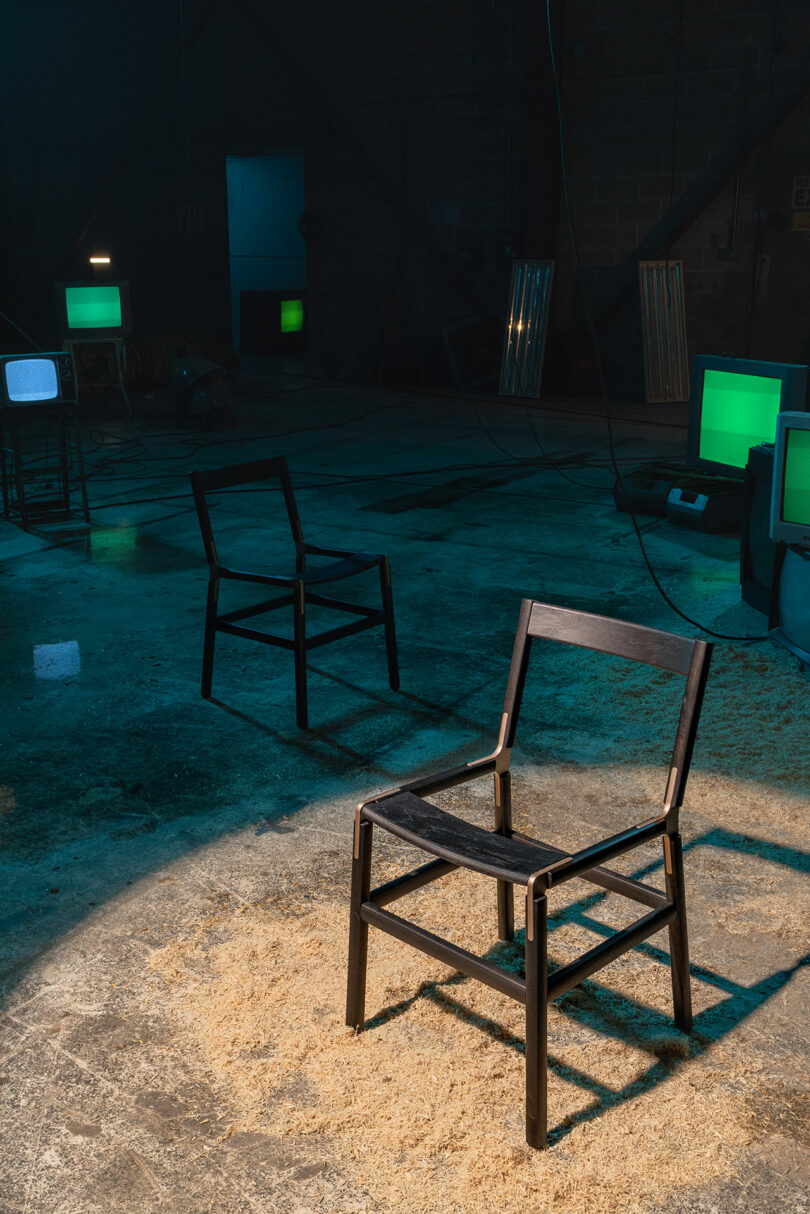 A black wooden chair sits in a spotlight on a rough concrete floor, with two more chairs, green-lit TV screens, and scattered sawdust in the background.