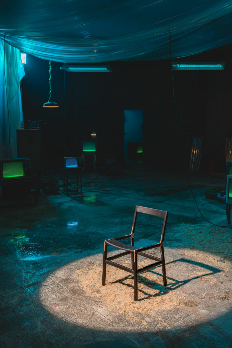 A single wooden chair is placed under a spotlight in a dark, industrial room with scattered sawdust and several old televisions emitting green light.