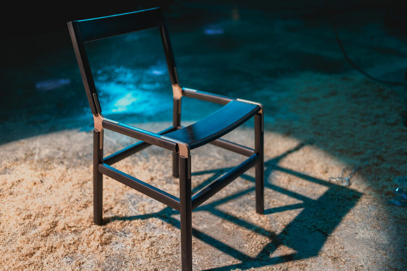 A modern black wooden chair with a minimalist design is placed on a textured, sawdust-covered floor under blue lighting.