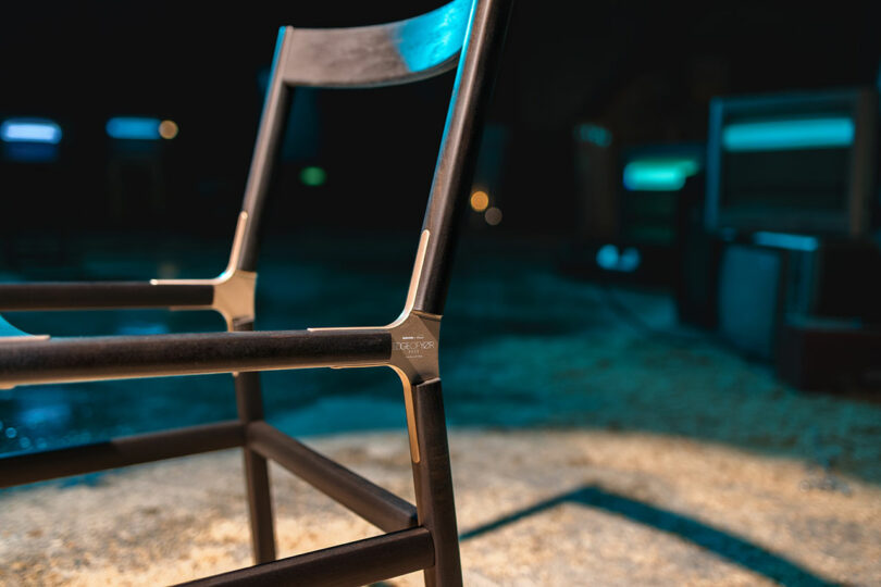 Close-up of a modern black chair with metal corner joints, set against a blurred background with blue and teal lighting.