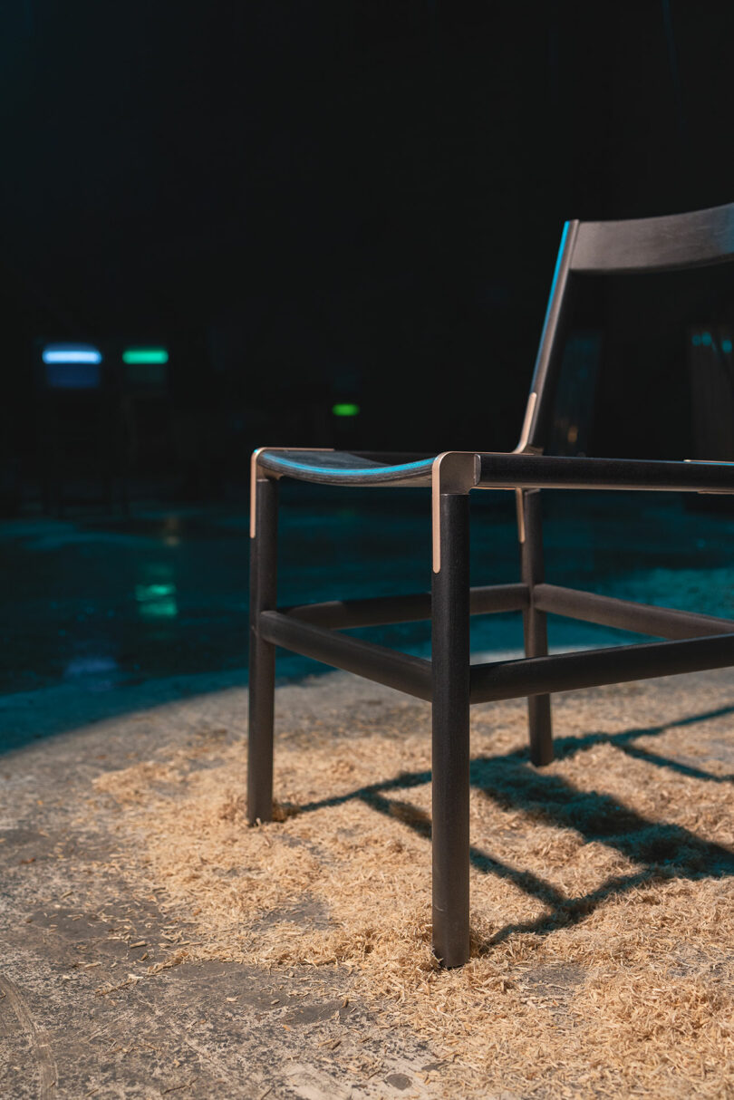 A black wooden chair sits on a concrete floor covered with sawdust, under low lighting with a dark, blurry background.