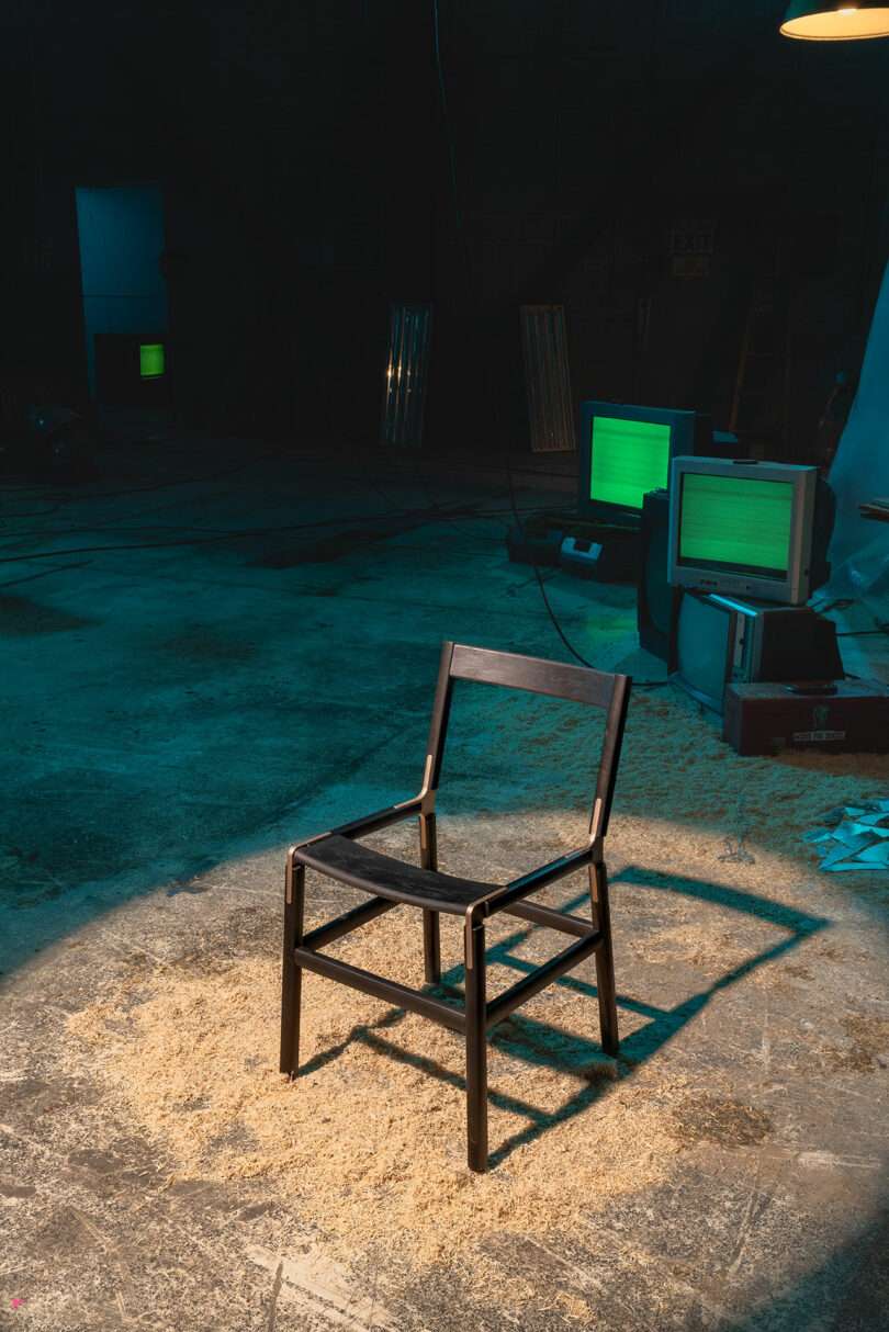 A wooden chair stands on a sawdust-covered floor in a dimly lit room with old computer monitors in the background.