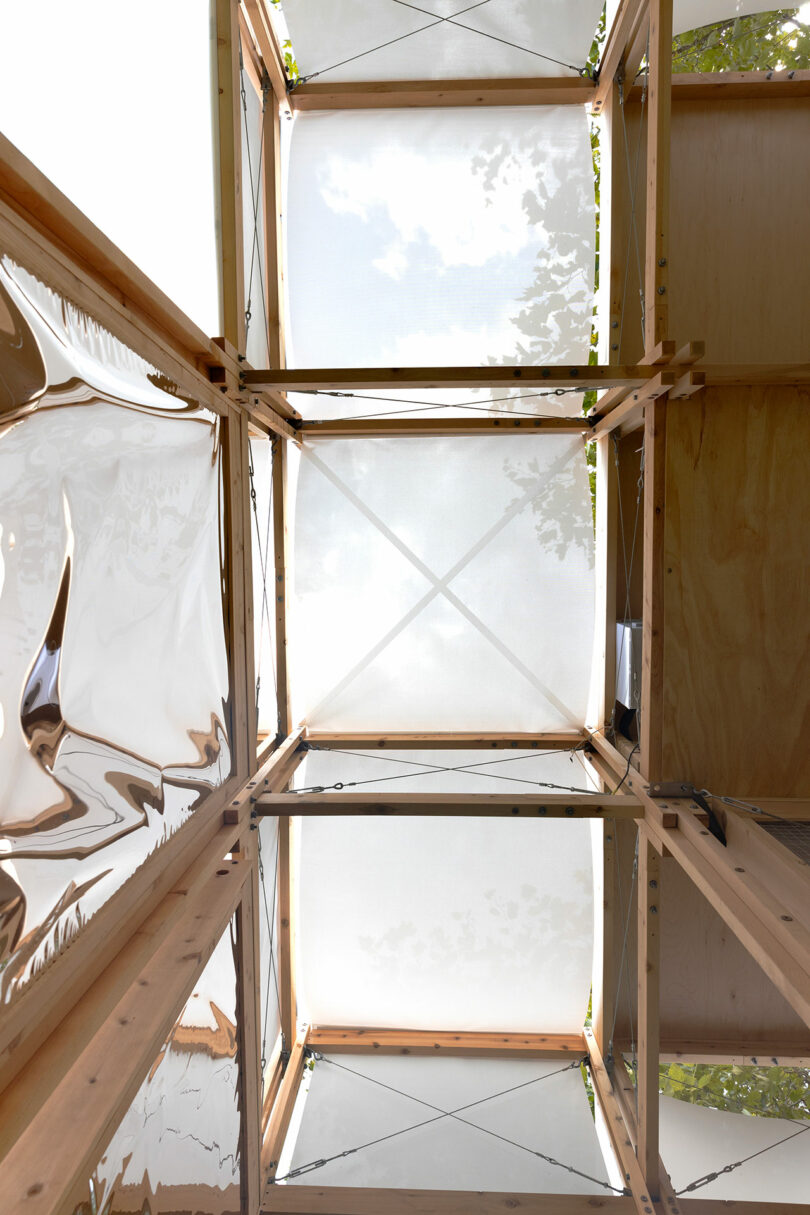 View looking up through a structure made of wood beams, translucent fabric panels, and reflective surfaces, with partial tree branches visible above.