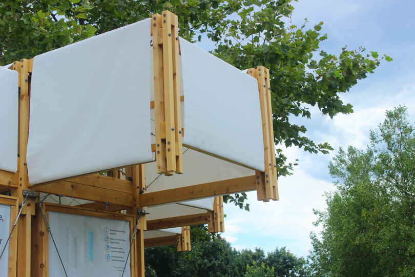 Close-up of a wooden outdoor structure with white fabric panels, set against trees and a cloudy sky.