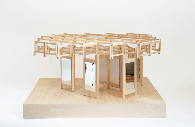 A wooden architectural model featuring a grid of interconnected frames on top and reflective panels on vertical supports, displayed on a light wooden base.