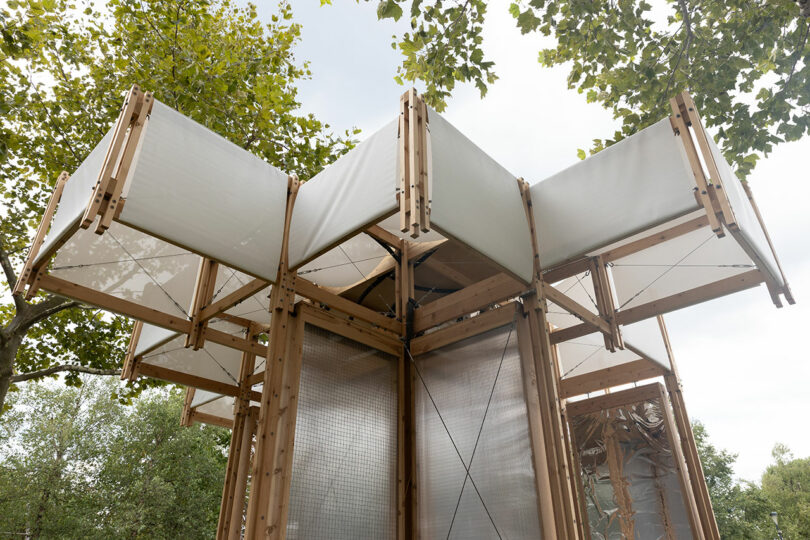 A wooden pavilion structure with large white fabric panels extends outward, surrounded by green trees and set against a cloudy sky.