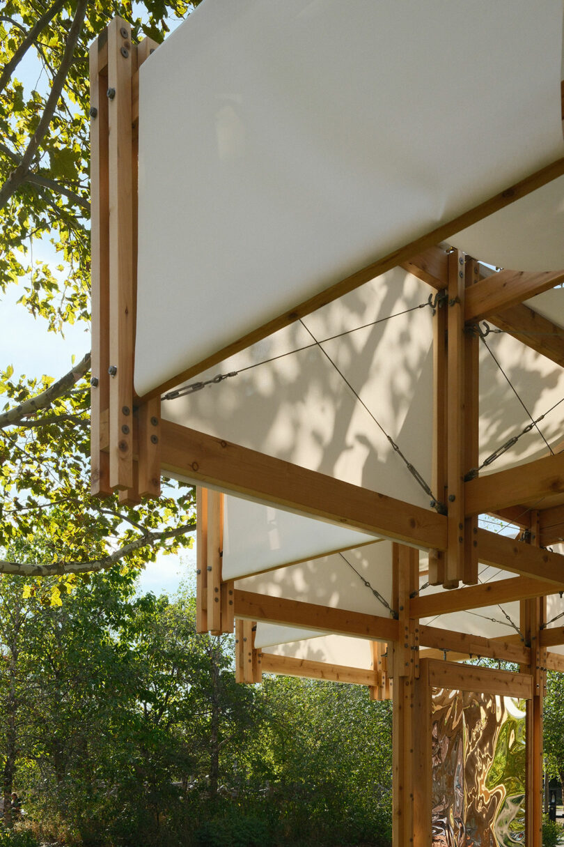 Close-up view of a wooden frame structure with white fabric panels and metal cables, set outdoors among green trees.
