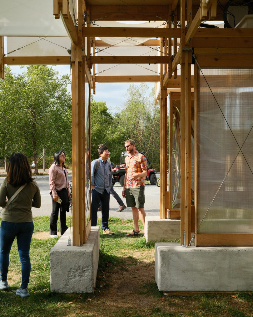 Four people stand and converse near a wooden and glass outdoor structure, with trees and a parked vehicle visible in the background.
