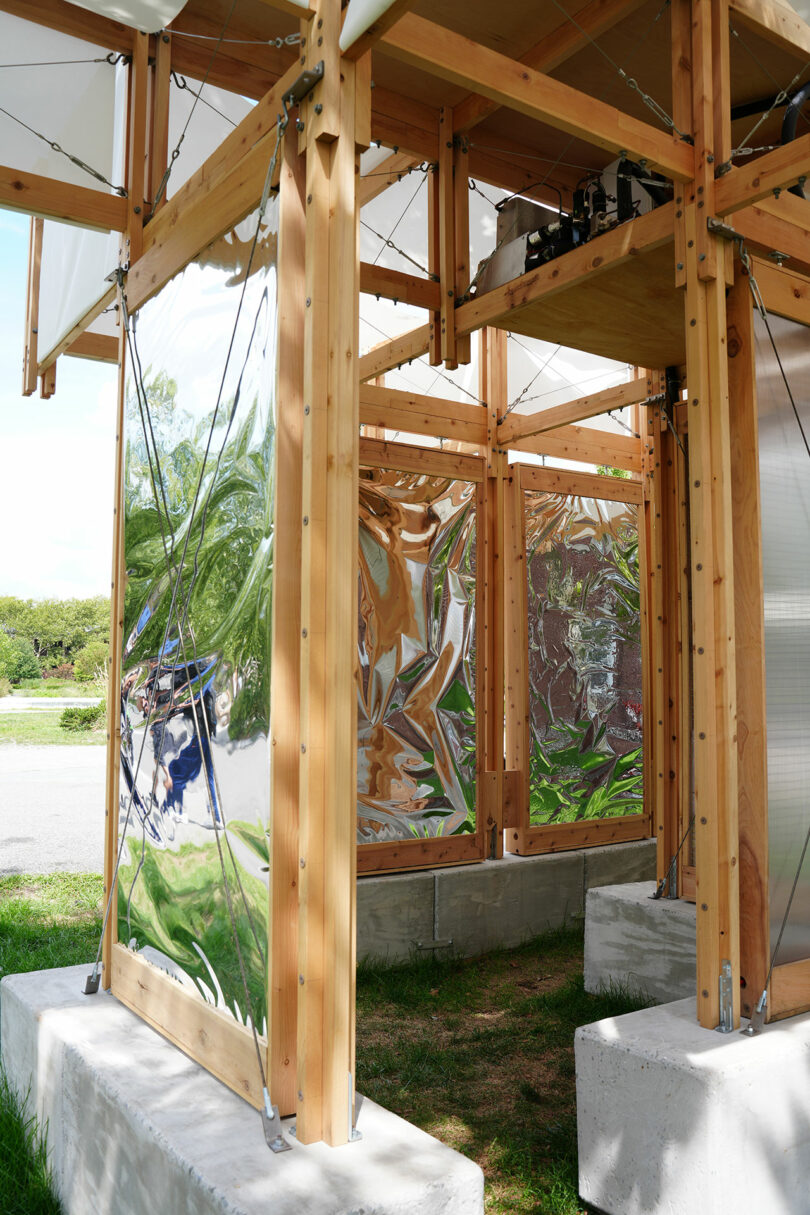 A wooden-framed outdoor structure with transparent and reflective metal panels, set on concrete bases, stands on grass near a paved area.