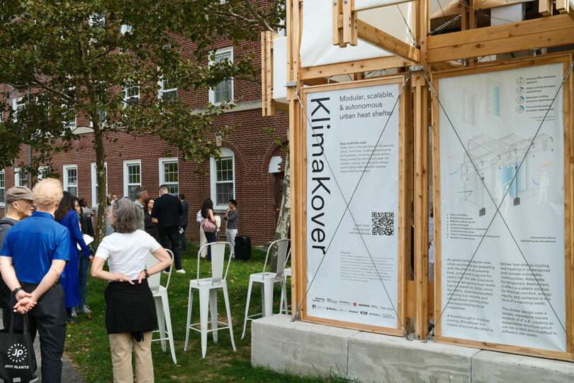 People gather outdoors near a brick building, viewing an exhibit titled 