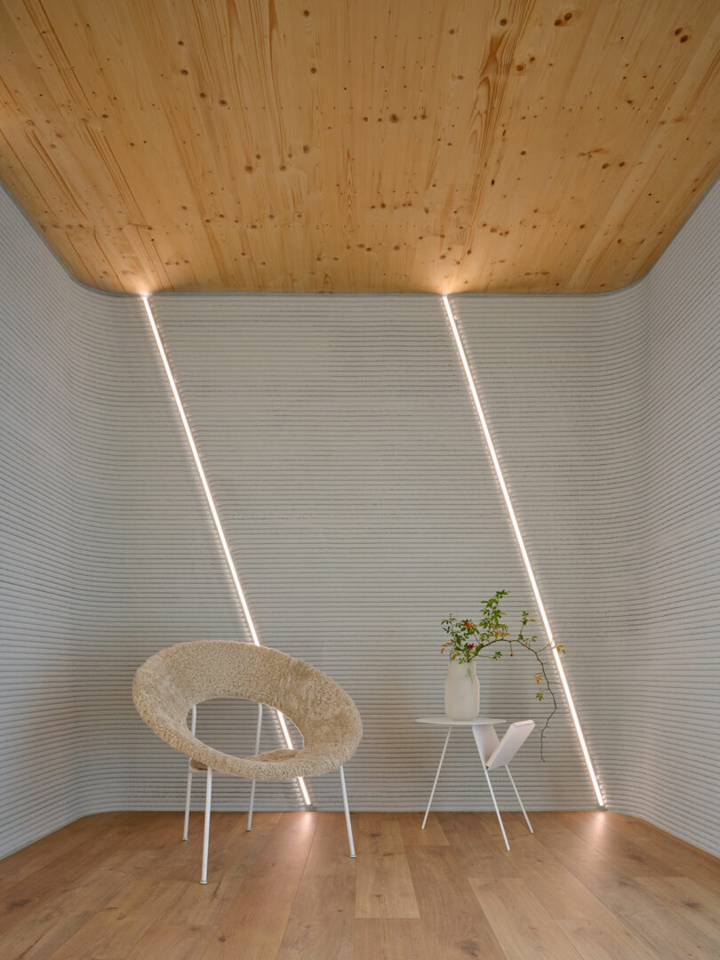 A modern room with a wooden ceiling, textured gray walls with diagonal LED lights, a woven chair, and a white side table holding a vase with branches.