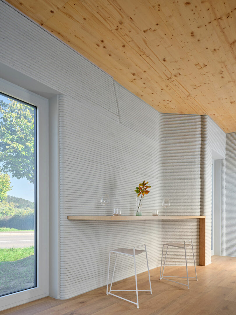 Minimalist interior with light wooden ceiling and floor, ribbed white wall, a narrow wooden counter, two white stools, and a single vase with flowers beside a large window.